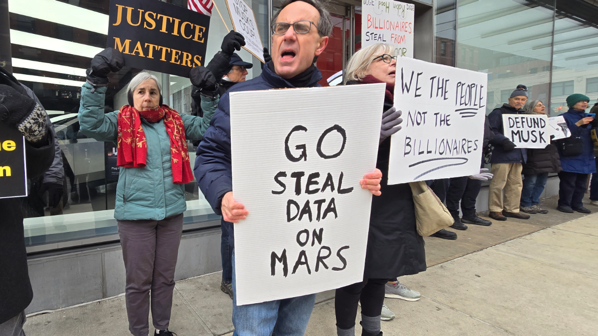 Man holds a "Go Steal Data on Mars" sign next to someone holding a "We the People, not the Billionaires" sign on a sidewalk.