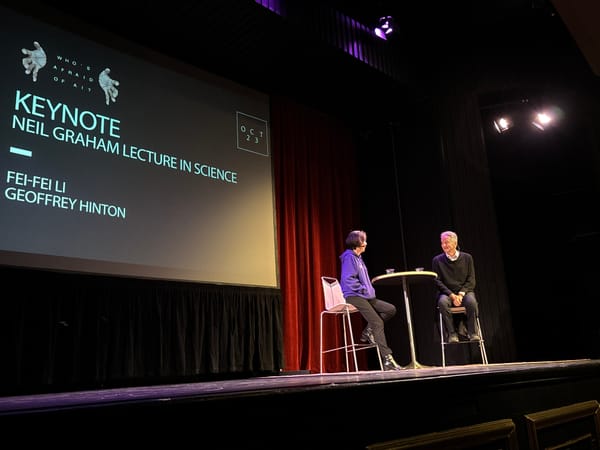 Two people seated at a table on a stage. The woman, Fei-Fei, wears purple and has short black hair. The man, Geoffrey, sits across from her in a black sweater and jeans. 